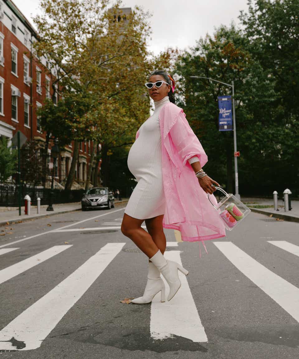 Hananah Fallis Bronfman walks across the street in New York. She is wearing white sunglasses, a white sweater dress, and a pink coat and carrying a clear bag with Poppi drinks inside.