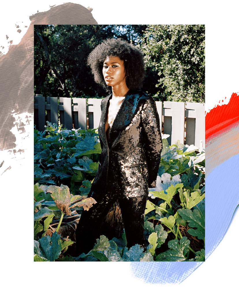 Amaya January model standing in a garden. She is wearing a black sequin blazer and has brown curly hair and a black cat eye makeup look. A brown, orange, red, and blue paint swatch is illustrated beneath the image.