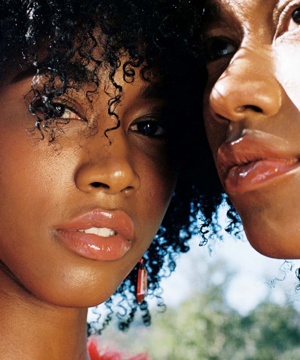 Twin models Alana and Amaya January stand face to face. One is staring softly into the camera with her brown curls falling in her face and has pink crystal earrings.