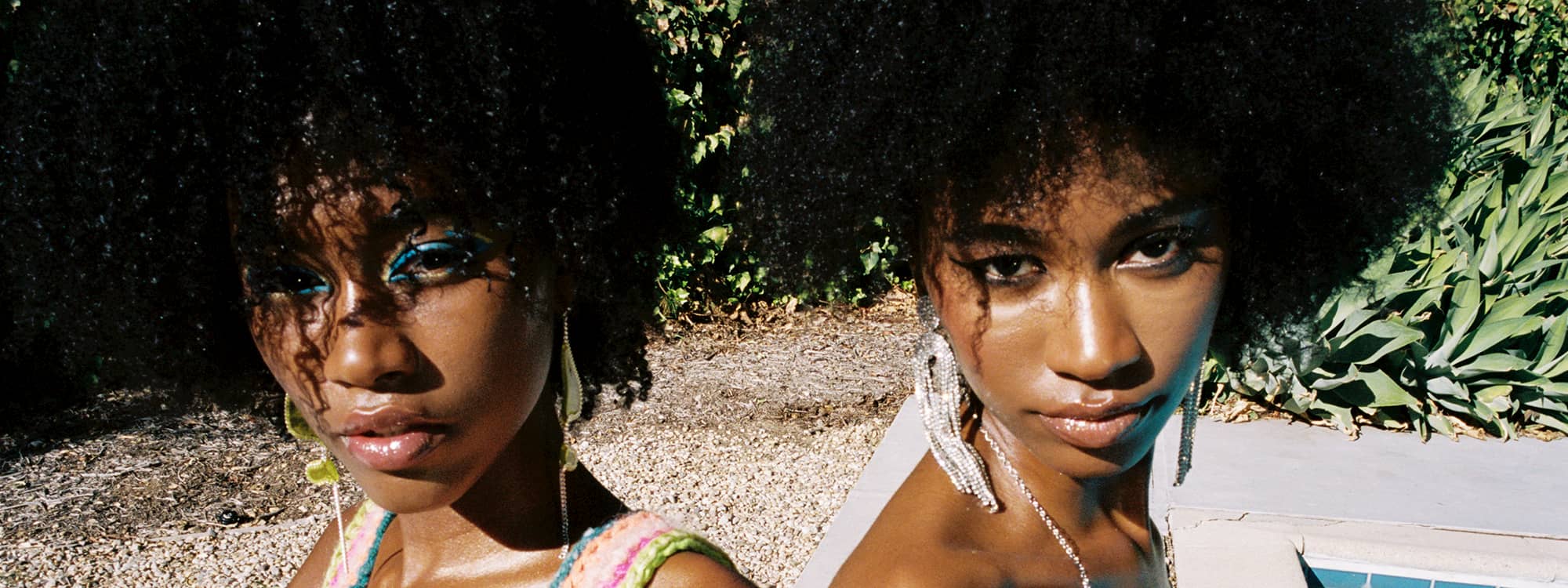 Twin models Alana and Amaya January, are sitting next to each other outside. One model is wearing bright blue eyeliner and has yellow earrings and a crochet colorful pink top. The other is wearing silver drop earrings and a silver necklace. Both have curly brown hair.