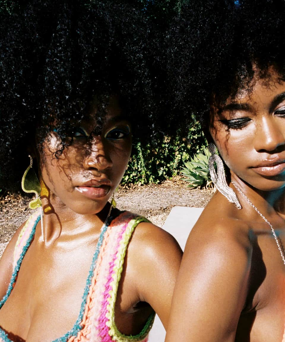 Twin models Alana and Amaya January, are sitting next to each other outside. One model is wearing bright blue eyeliner and has yellow earrings and a crochet colorful pink top. The other is wearing silver drop earrings and a silver necklace. Both have curly brown hair.