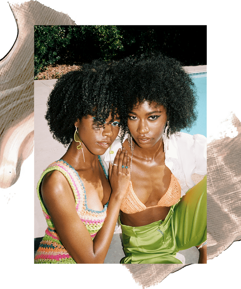 Model Amaya January stands outside in a peach crochet bikini top and silver earrings. She has brown curly hair and is standing in front of trees and a bright blue sky. In the second image she is seated next to her sister Alana January and wearing green silk pants and a white button-down shirt that is open to reveal the bikini top. Her sister is wearing a pink and green crochet dress. There is an illustration of brown and peach paint behind them.