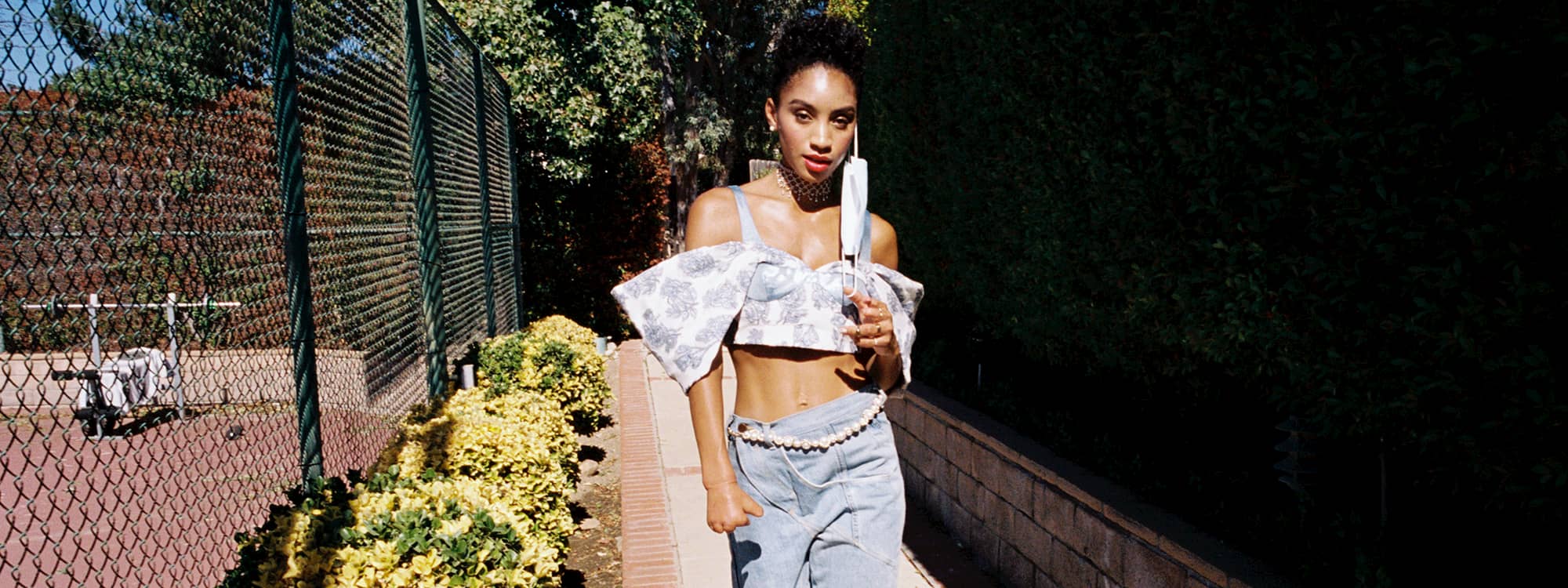 Lyric Mariah walks down a pathway near a fence. She is wearing a cropped jean top and jean pants with a pearl belt. She is taking her face mask off.