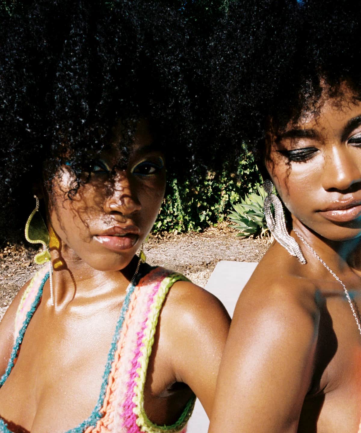 Twin models, Alana and Amaya January. They have brown curly hair and are standing shoulder to shoulder. One is wearing a crochet top and the other has a bikini top on. Their skin is glowing in the sunlight.