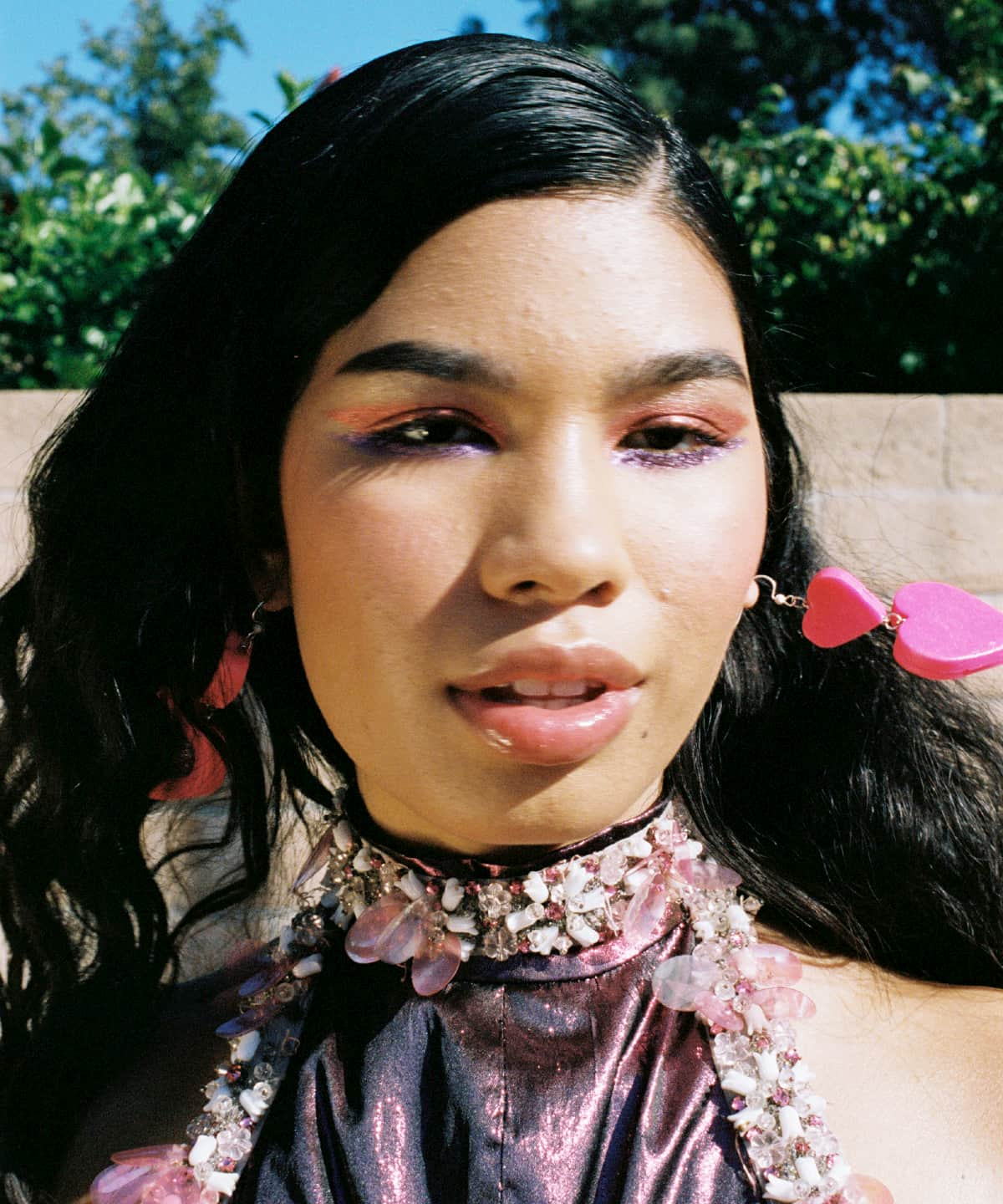 Model Ariel Toole wearing a high-neck purple top with soft crystal appliques. She is wearing purple and orange eyeshadow, and has bright pink heart-shaped earrings.