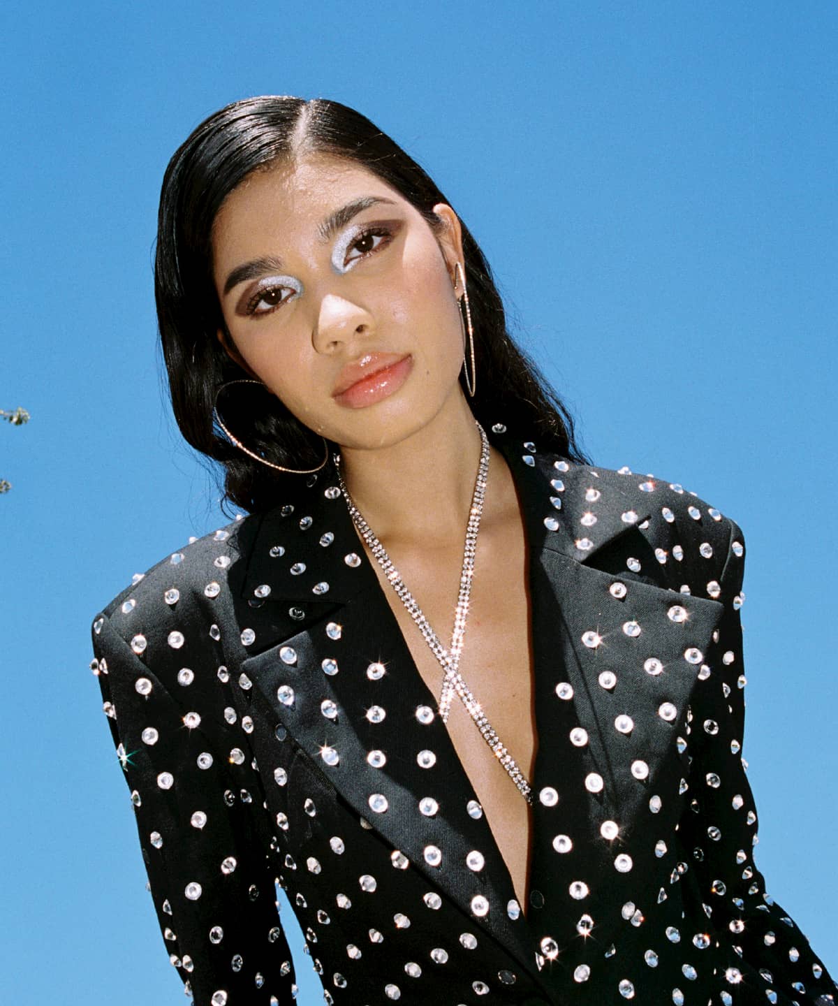 Model Ariel Toole outside with long brown hair and silver eyeshadow. She is wearing a black blazer studded with silver crystals and a silver necklace and hoops.