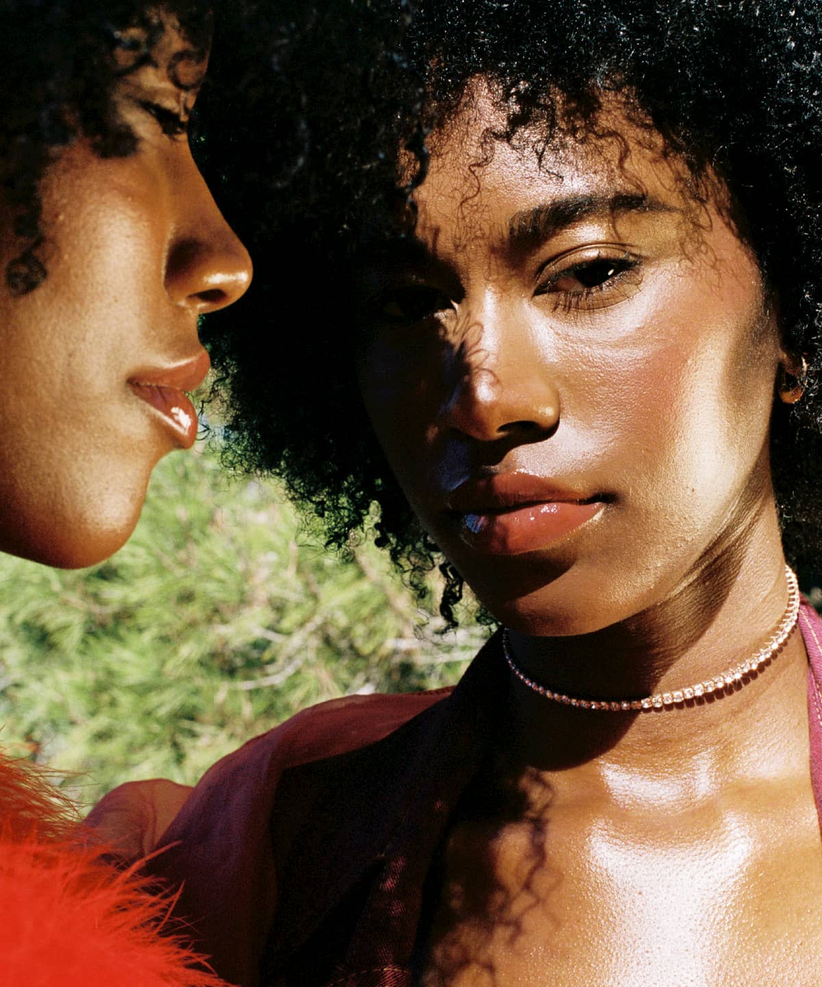 Close-up of twins models, Alana and Amaya January, with curly hair standing next to each other. One is wearing a red feather top and the other is wearing a pink top. Their skin is glowing in the sunshine.