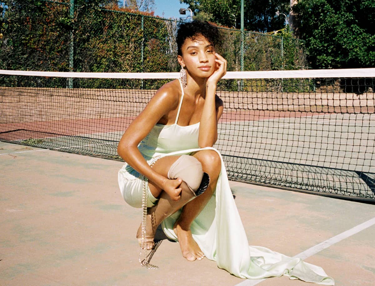 Model Lyric Mariah wearing a long green silk dress and kneeling on a tennis court with her arm wrapped around her prosthetic leg. She has curly brown hair and her makeup is soft and glam.