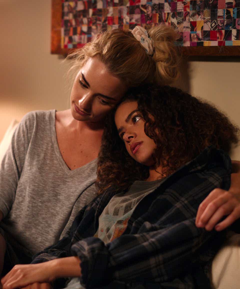 Brianne Howey and Antonia Gentry appear as Georgia and Ginny. They are sitting on Ginny's bed and Georgia is comforting her daughter. She wears a grey v-neck sweater and a floral scrunchie. Ginny is wearing a plaid shirt over a t-shirt. 