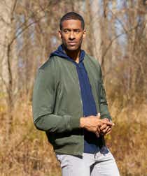 Matt James appears on The Bachelor. He is standing in the woods outside Nemacolin Resort and wearing a green windbreaker over a navy blue hoodie with grey jeans.