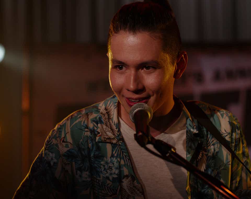 Mason Temple appears as Hunter in Ginny &amp; Georgia. He is onstage singing into a microphone. He's wearing a blue and white Hawaiian shirt over a white t-shirt. 
