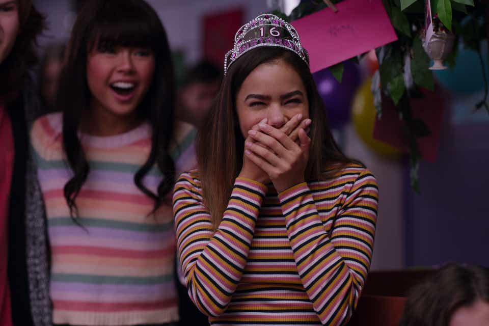 Antonia Gentry appears as Ginny in Ginny and Georgia. She is being surprised at her 16th birthday party, so she's laughing and covering her mouth. She is wearing a multi-colored striped shirt with long sleeves and a pink and silver crown with the number 16 on it. 