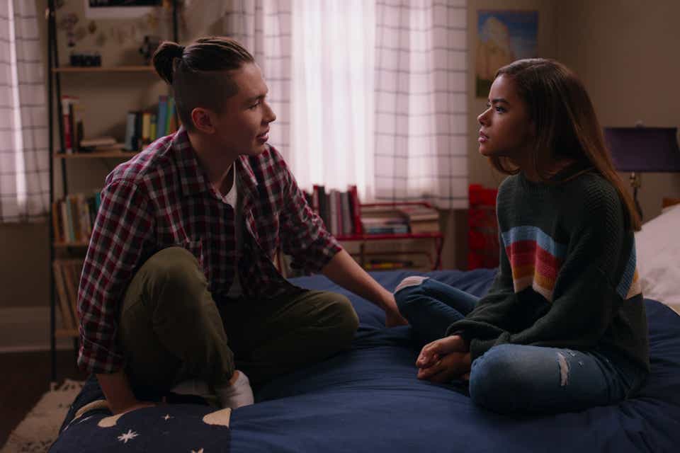 Mason Temple and Antonia Gentry appear in Ginny and Georgia. He is wearing his hair in a small bun and is dressed in a red plaid shirt and green pants. She is wearing a navy blue sweater with rainbow stripes across the middle and a pair of ripped jeans. Her usually curly hair is straightened. Both actors are sitting on a bed facing each other, deep in conversation.