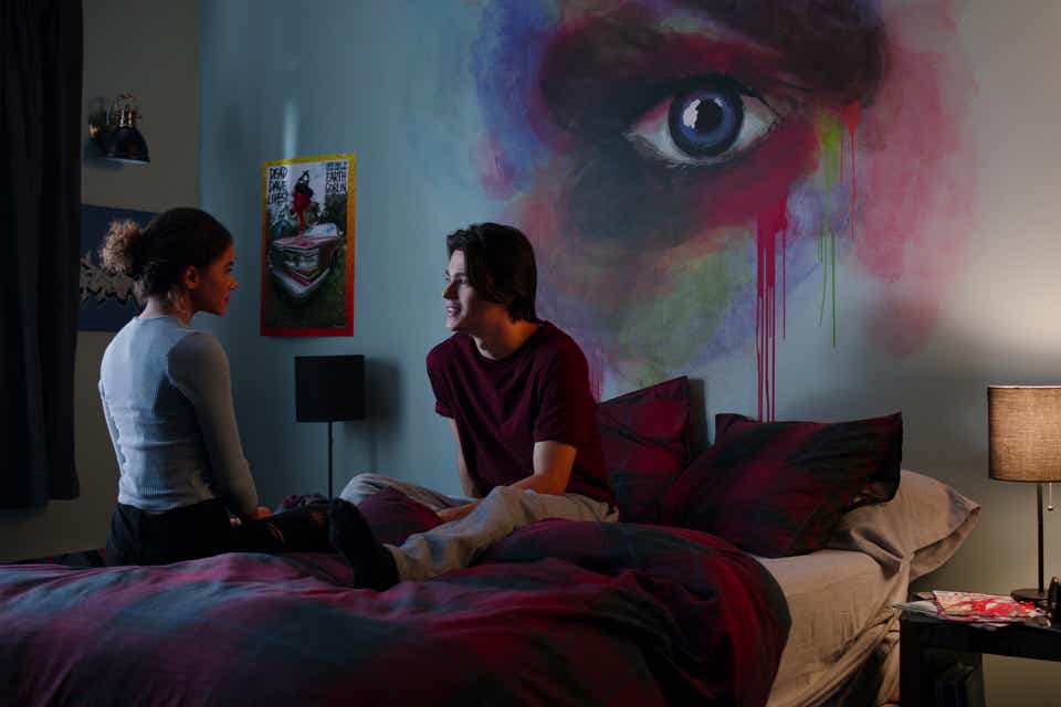 Antonia Gentry and Felix Mallard appear in Ginny and Georgia. He is wearing a red shirt and grey pants. She is wearing a light blue sweater. Her hair is in a bun. Both actors are sitting on a bed facing each other, deep in conversation.