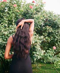 Black woman with long brown hair in a black dress stands outside among tall bushes with her back turned to the camera