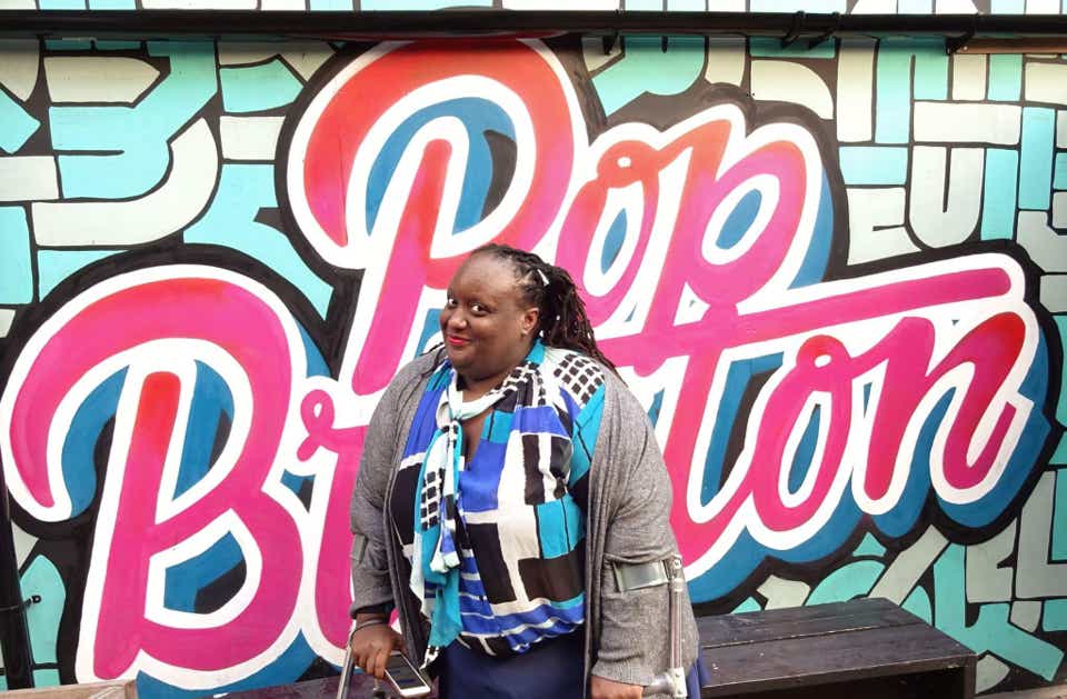 Imani Barbarin, a Black woman with Black locs in a ponytail down her back, stands on crutches in a multi-colored Blue and white shirt, smiling, in front of a Blue tiled wall with pink ombre graffiti that reads "Pop Button"