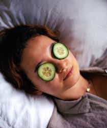 Person lies on pillow with cucumber slices on their eyes