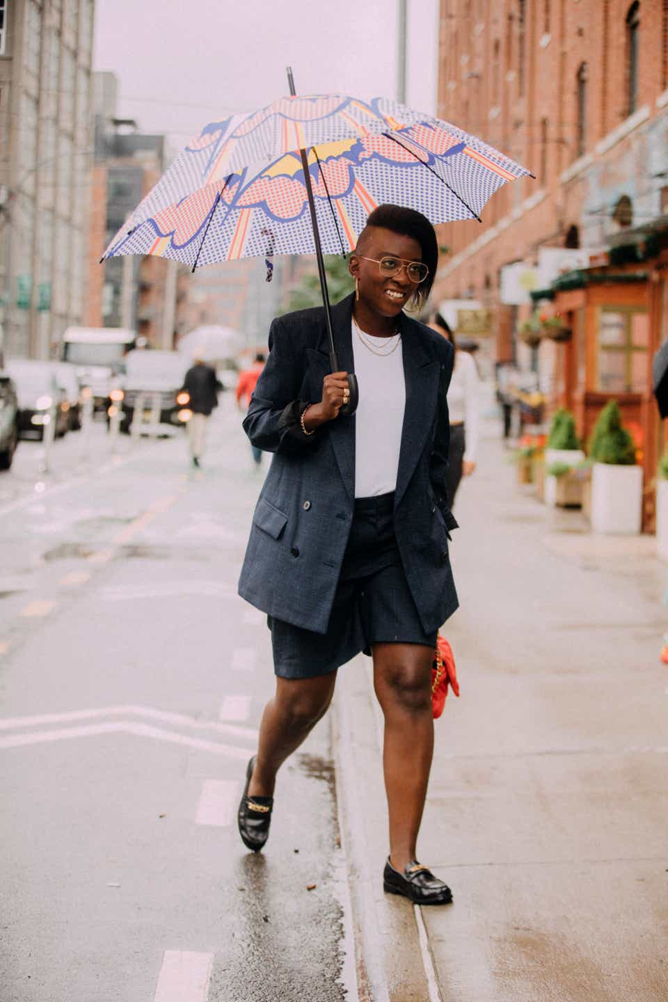 Nikki Ogunnaike wears a blazer, white top, and matching shorts and black loafers under an umbrella