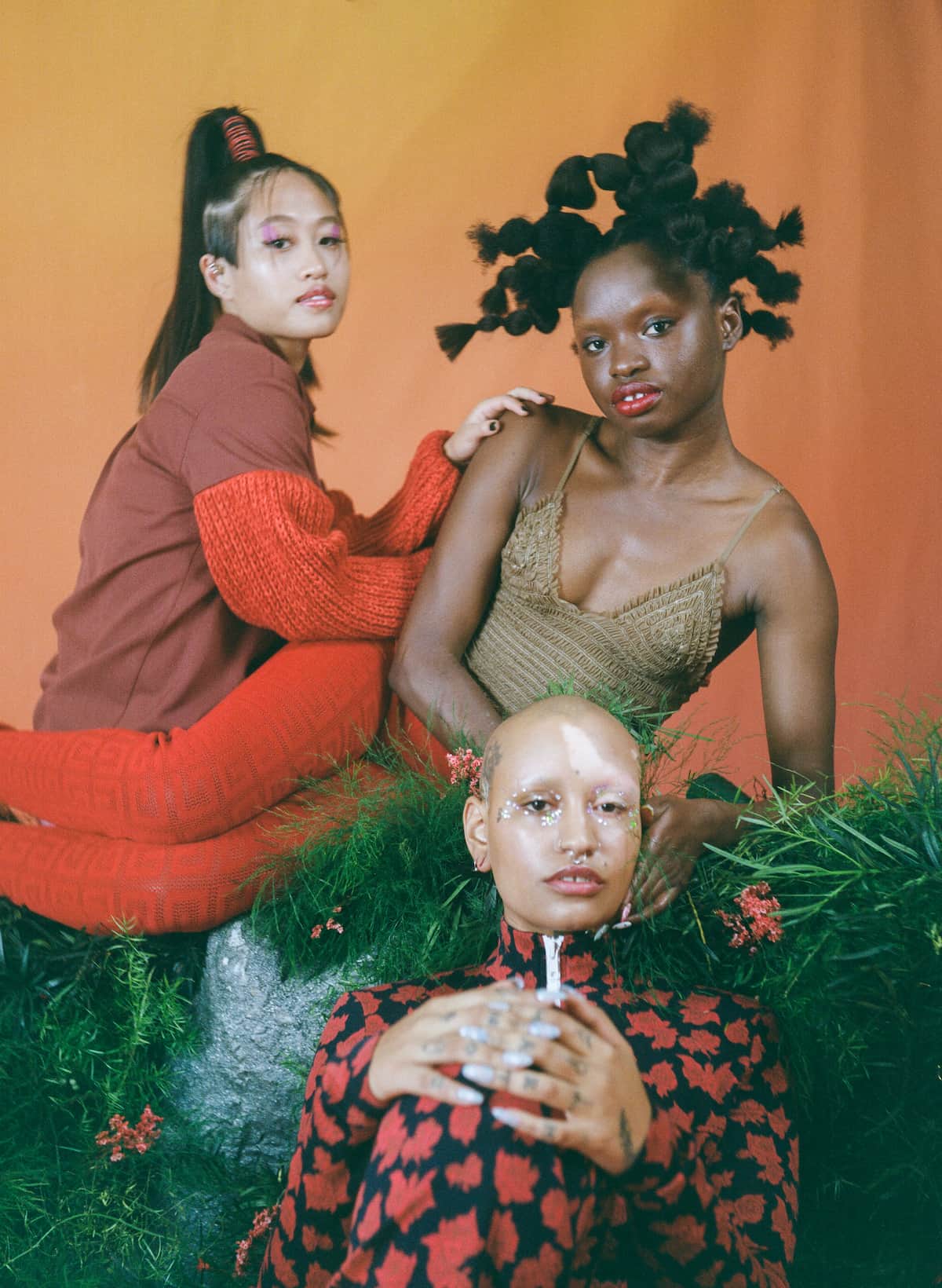 Three models seated in front of an orange-and-yellow background, wearing red ensembles. 