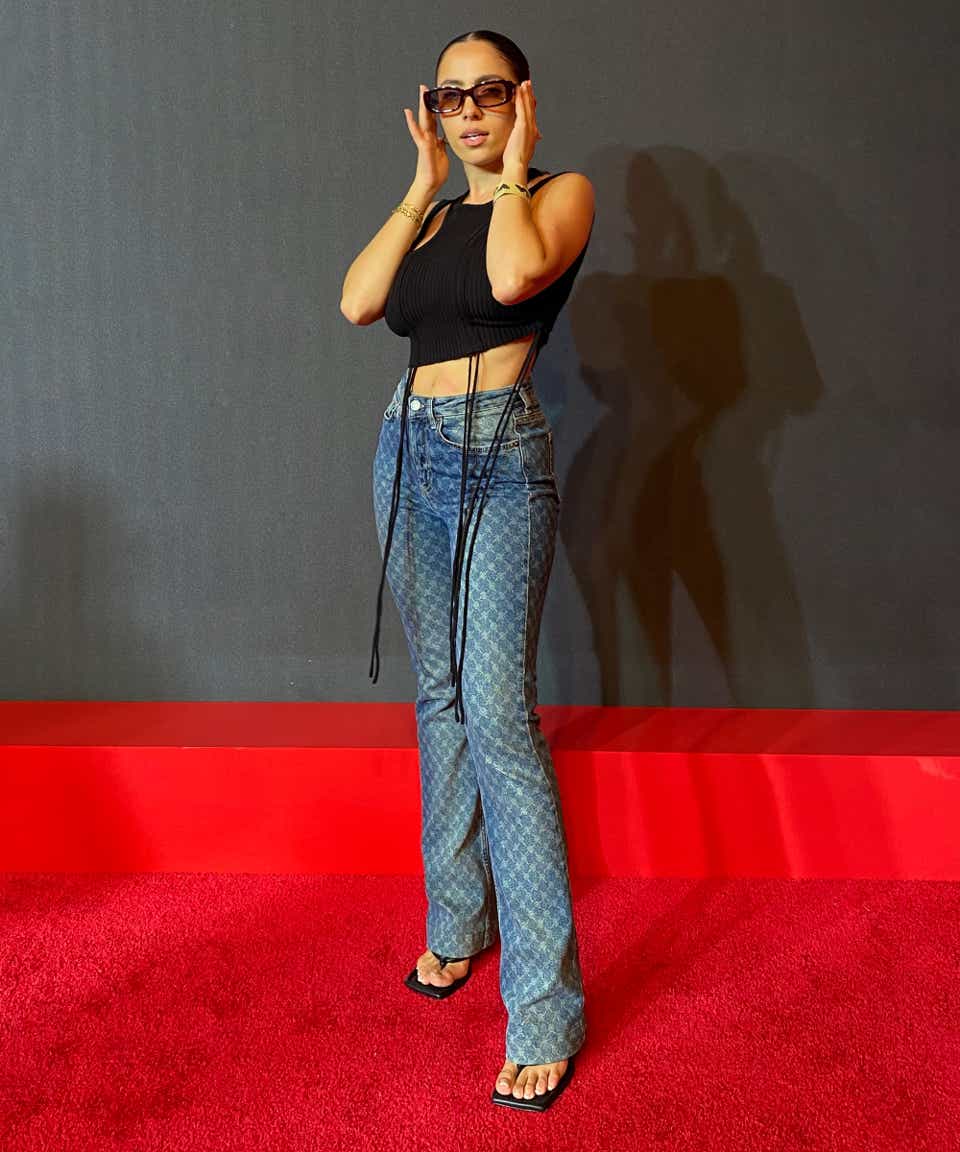a woman poses on the red carpet wearing sunglasses, a black cropped top, and blue jeans