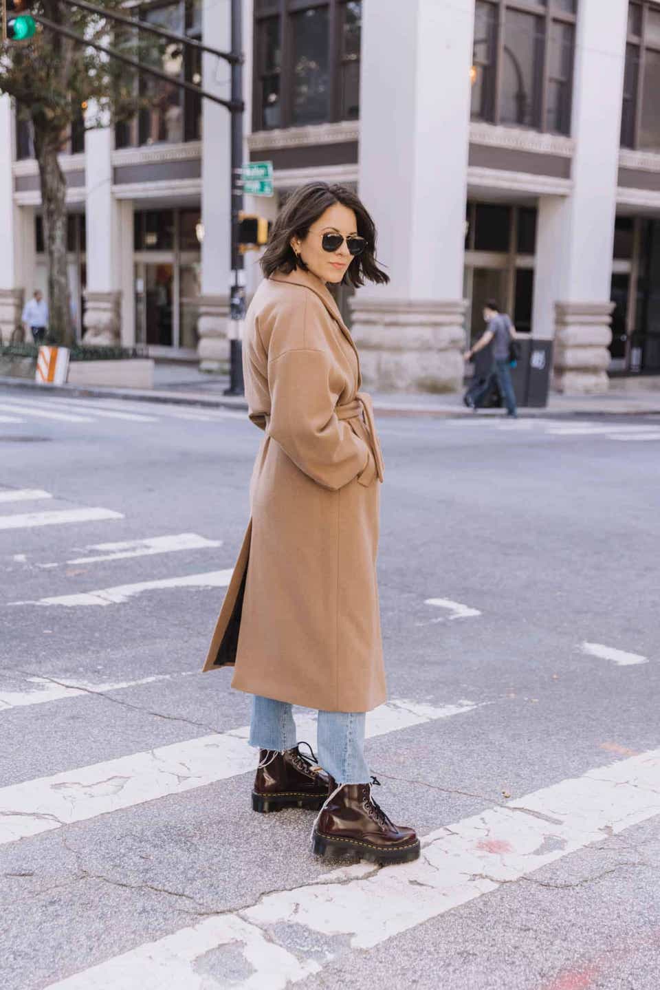a woman walks on the street wearing a camel coat, blue jeans, and red dr. martens boots