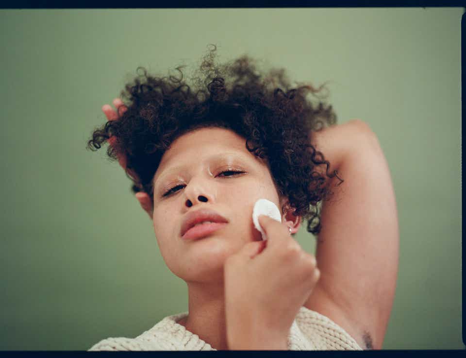 Model with curly hair holding a cotton round to their face photographed against a green backdrop