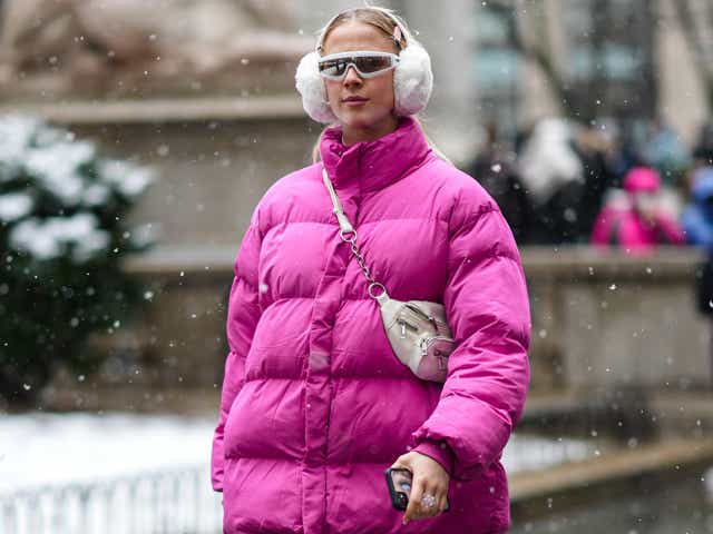 A guest wears pale pink hair clip, a white fluffy ear muffs, a pink shiny oversized high neck puffer jacket, black shiny leather large pants, a white leather fanny pack bag, black shiny leather ankle boot, outside Ulla Johnson