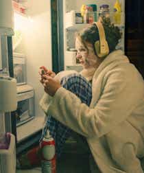 Person sitting in front of a fridge wearing a bath robe, face mask and headphones while using their phone