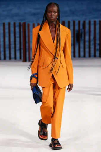 a model wears a bold orange two-piece suit, paired with a blue handbag, and a pair of black flats chunky sandals at the proenza schouler spring 2022 show