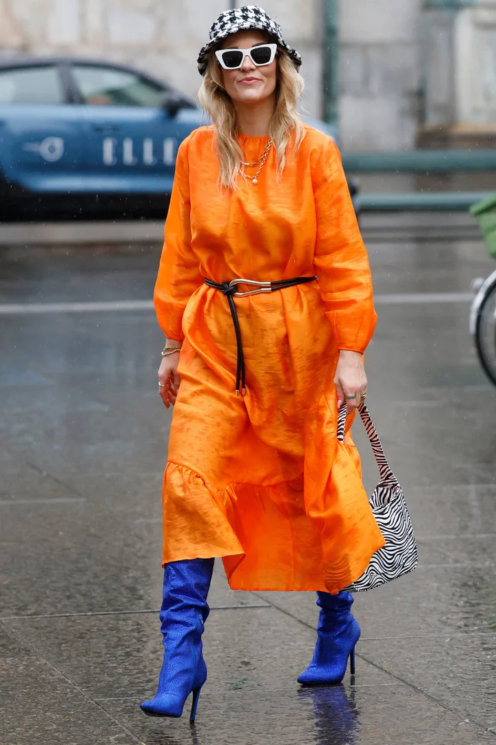 a woman wears a bold orange dress, paired with blue boots, a zebra print handbag, a black belt, white sunglasses, and a black and white bucket hat