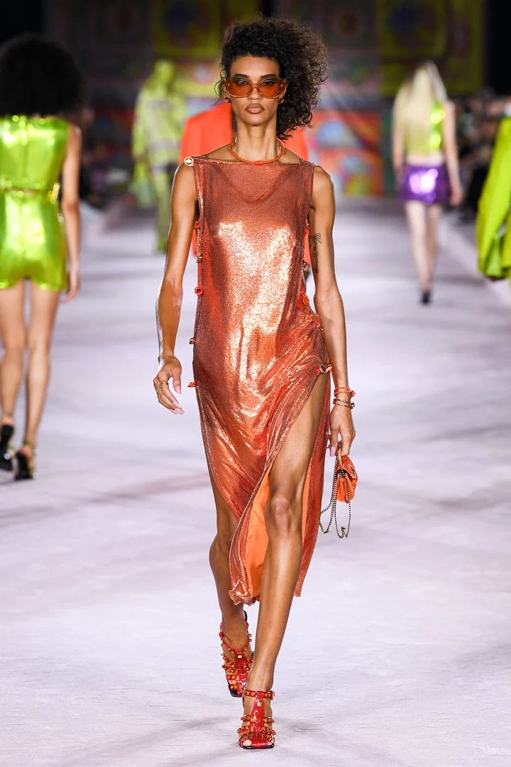 a model wears an orange metallic mesh dress, paired with orange studded heeled sandals, an orange mini handbag, and orange sunglasses at the versace spring 2022 show