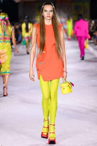 a model wears a bold orange mini dress, paired with neon green tights, pink platform heels and a yellow handbag at the versace spring 2022 show