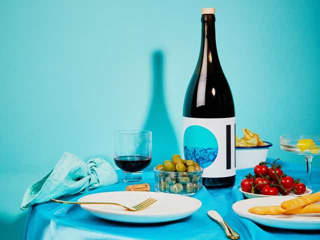 Dark green glass bottle of Citizens of Soil olive oil on blue table, with wine glasses, olives, tomatoes and plates.