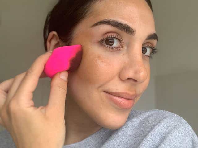Jacqueline holding a bottle of fake tan to camera and a pink beauty blender to her face