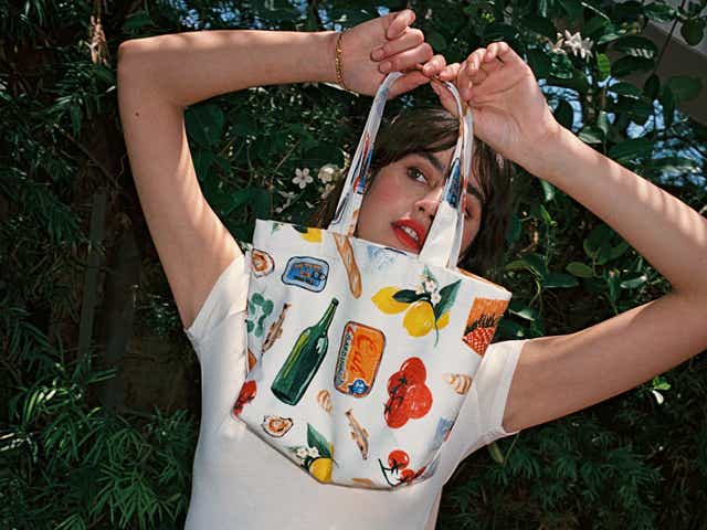 A model holding a white tote printed with lemons, tomatoes, bread, wine, and sardines.