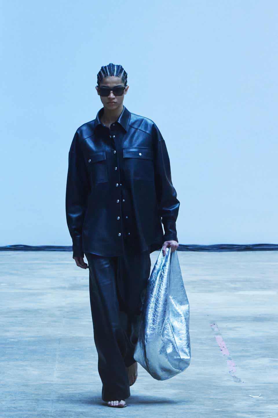 A runway model wearing a black leather blazer with matching pants and an oversized silver bag.