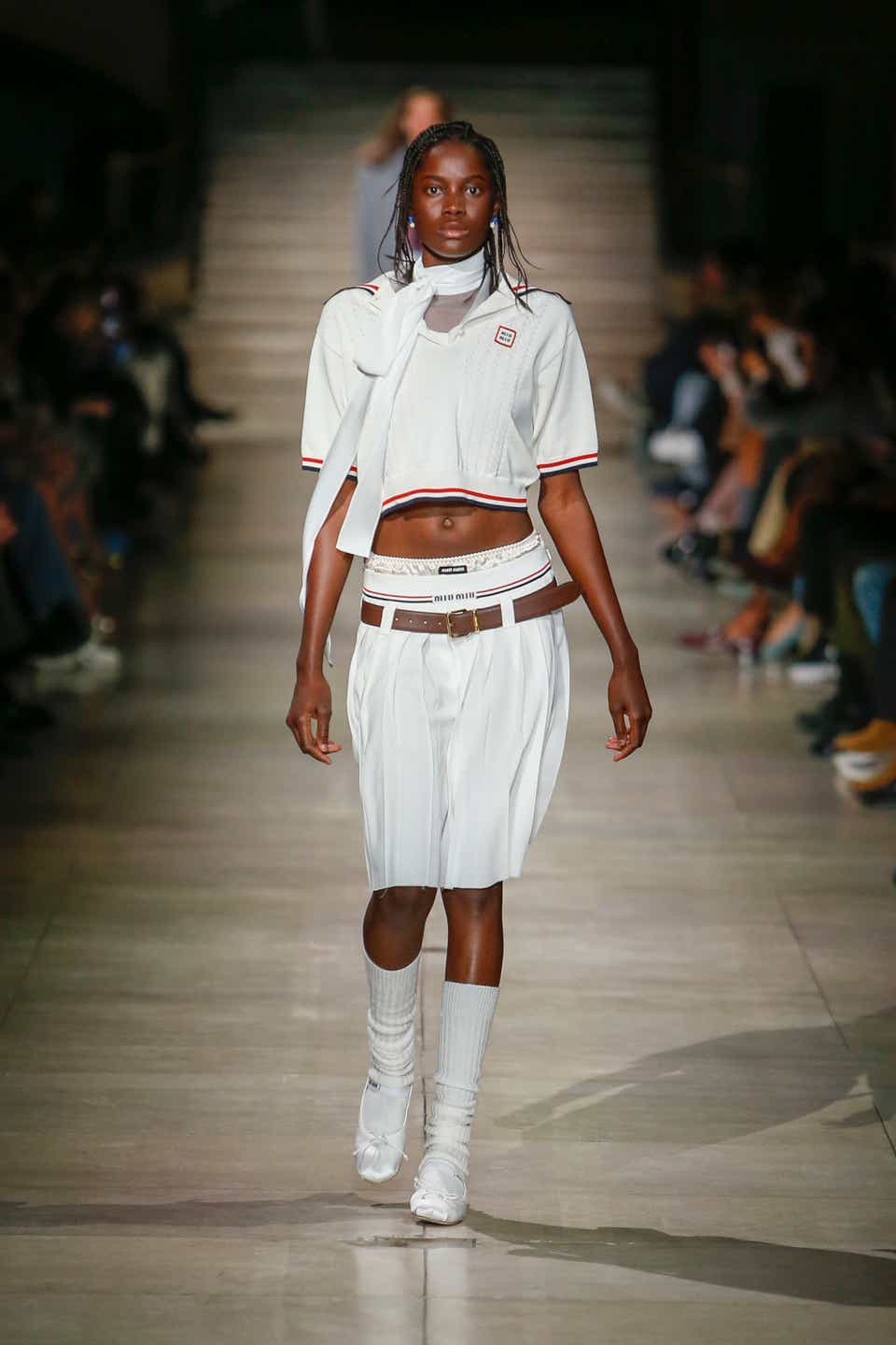A runway model wearing a white polo with a matching pleated skirt and high-knee socks.