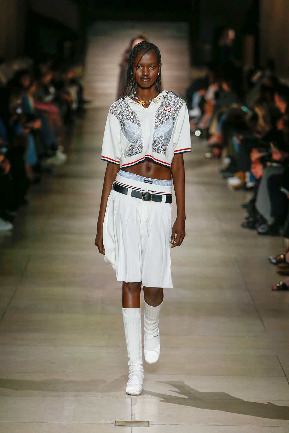 A model wearing a knee-length pleated white skirt on the Miu Miu Fall 2022 runway.