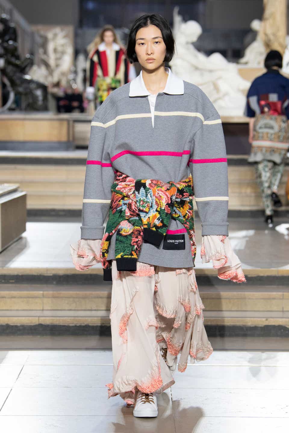 A model wearing a gray striped polo shirt with a floral sweater tied around the waist on the Louis Vuitton Fall 2022 runway.