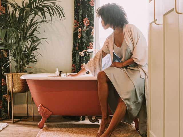 A beautiful woman perches on the side of an elegant red roll top bathtub, and waits for it to fill up. She wears a silky robe. Light floods through the window, backlighting the relaxing scene and giving it a dreamy vibe.