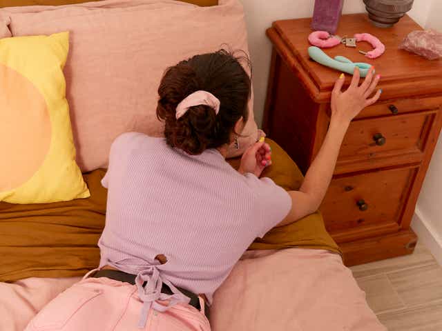 Woman on her stomach in bed reaching for a vibrator on her bedside table
