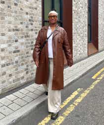 Ebony wearing a white shirt, brown leather trench coat, beige trousers and green boots