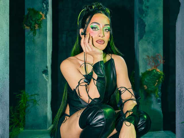 A photo of a light-skinned women bending over in heavy green makeup and a black leather outfit.