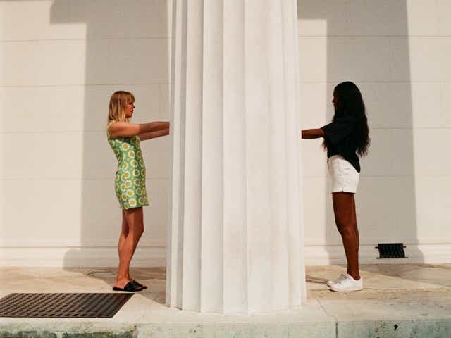 Two women standing on either side of a pillar reach out for each other